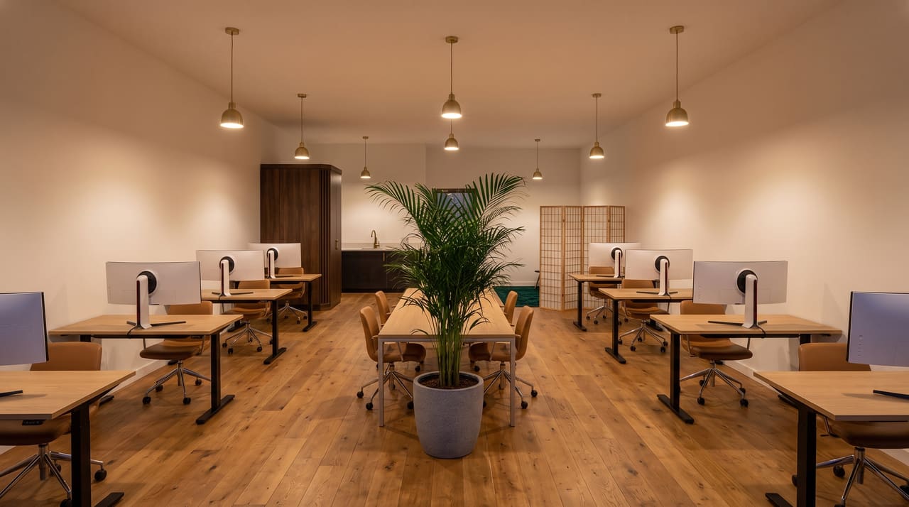 The sunlit main floor: light oak standing desks with 34-inch curved ultrawide monitors and cognac leather chairs against warm white walls.