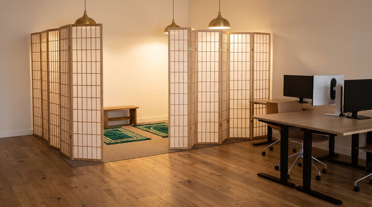 The open musalla corner: three emerald prayer rugs on the office carpet, an oak shoe bench, and a brass medallion on the warm white wall, with desks a few steps away.
